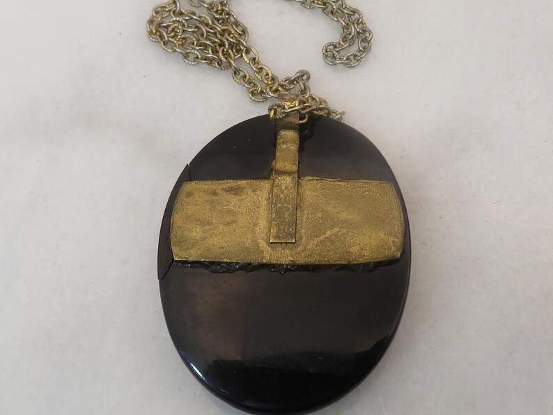 May include: A vintage necklace featuring a large, oval black pendant with a gold-colored metal overlay. The pendant hangs from a silver-colored chain. The gold metal has a rectangular design.