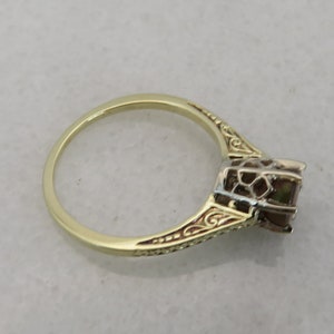 May include: A gold ring with an ornate design. The ring features a square-cut gemstone and intricate detailing along the band. The ring is set against a neutral background, highlighting its elegant design.