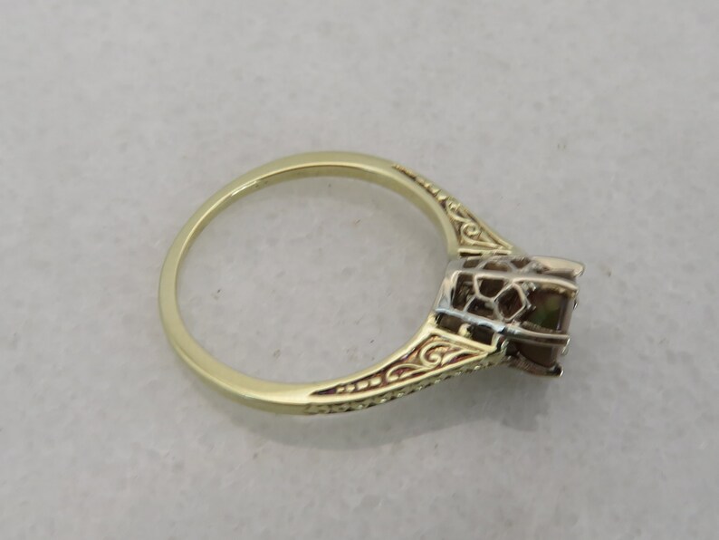 May include: A gold ring with an ornate design. The ring features a square-cut gemstone and intricate detailing along the band. The ring is set against a neutral background, highlighting its elegant design.
