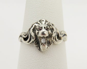 May include: A silver ring with a detailed lion head design. The lion has red eyes and a single clear stone in its mouth.