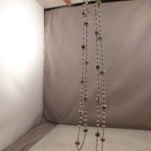 May include: A long silver chain necklace with white and brown beads. The necklace is hanging from a hand.