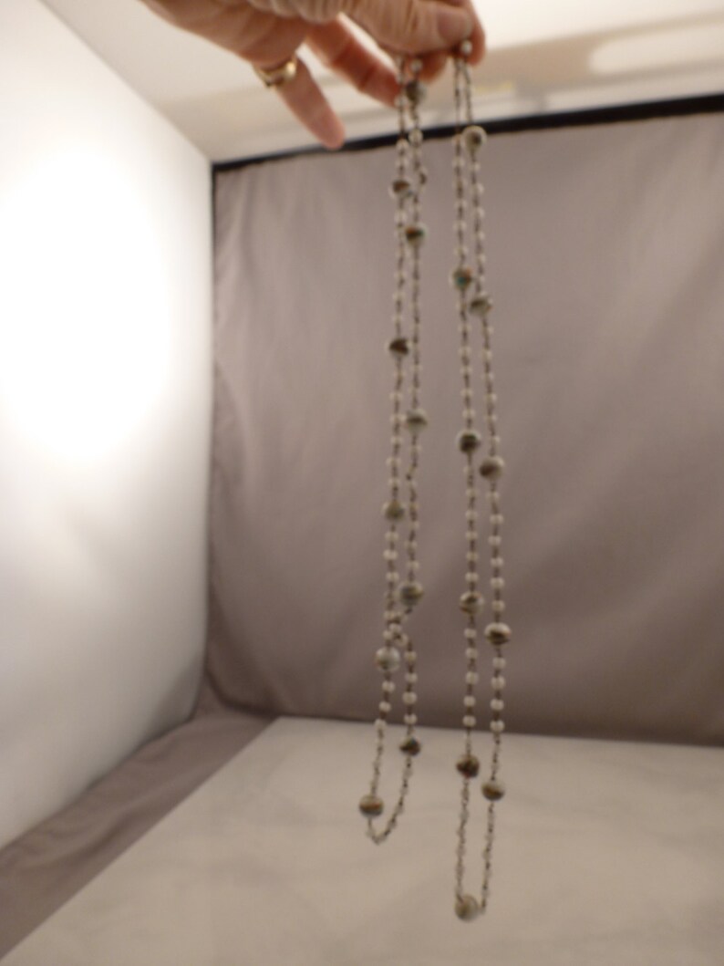 May include: A long silver chain necklace with white and brown beads. The necklace is hanging from a hand.