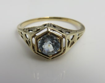May include: A gold-toned ring with an oval, light blue gemstone set in a filigree design. The ring's setting features an openwork geometric pattern, creating an intricate and delicate appearance. The ring is likely a vintage or antique piece.
