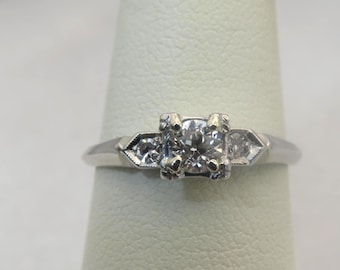 May include: A silver engagement ring with a square-cut center diamond and two smaller diamonds on either side. The ring is set on a white background.