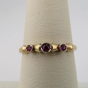 May include: A gold ring featuring three round, deep pink rubies set in a row. The center ruby is slightly larger than the two flanking stones, and each is held in a simple metal setting integrated into the smooth, polished gold band.