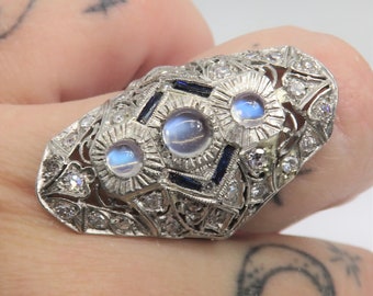 May include: A silver ring with three moonstone gemstones and smaller diamonds set in a filigree design. The ring also features blue sapphires.