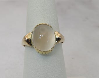 May include: A gold ring with a clear oval-shaped gemstone set in a bezel setting. The ring is on a white display stand.