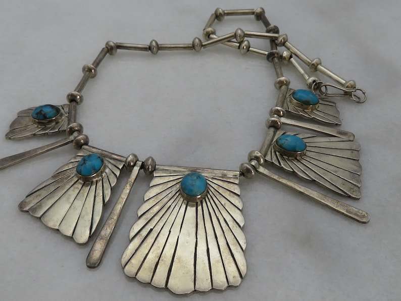 May include: A silver necklace with turquoise stones and a fan-shaped design. The necklace is made of sterling silver and features a unique geometric pattern.