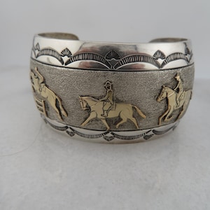 May include: Silver cuff bracelet with gold-colored horse and rider figures. The bracelet has a textured center band and decorative edges. The design features three horse and rider figures around the bracelet.