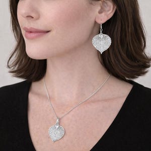 May include: A silver-toned jewelry set consisting of a pendant necklace and matching drop earrings. Each piece features a delicate, intricate leaf-shaped design with a fine, openwork filigree pattern that mimics the veins of a natural leaf.