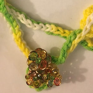 May include: A multi-colored, jeweled flower charm with green, pink, and yellow stones. The charm is attached to a braided cord with green, white, and yellow strands.