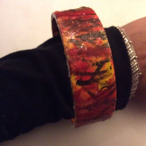 May include: A colorful, hand-painted bangle bracelet with red, orange, yellow, and black abstract designs. The bracelet is made of a thick, durable material and has a smooth, glossy finish.