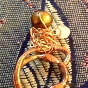 May include: A handmade copper-colored ring with a textured surface, featuring a small gold-colored bead and a white bead. The ring is set against a patterned blue and white background. Jewelry.