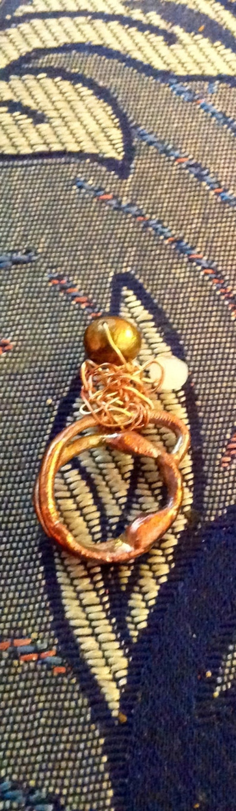 May include: A handmade copper-colored ring with a textured surface, featuring a small gold-colored bead and a white bead. The ring is set against a patterned blue and white background. Jewelry.