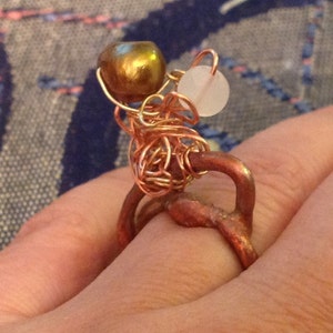 May include: A handmade copper ring featuring a cluster of copper wire, a golden-colored bead, and a small, round, white bead. The ring has a unique, artistic design with a rustic, organic aesthetic. The ring is worn on a finger.