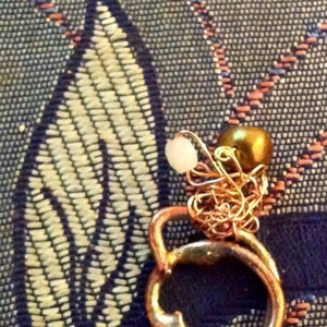 May include: A copper-colored ring with a small gold-colored bead and a white bead attached by a delicate chain. The ring is set against a patterned blue and beige fabric.