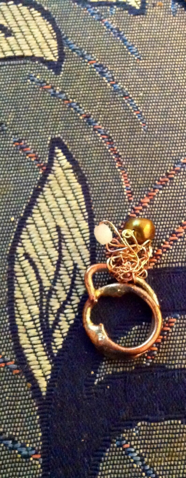 May include: A copper-colored ring with a small gold-colored bead and a white bead attached by a delicate chain. The ring is set against a patterned blue and beige fabric.