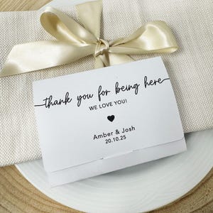K&ouml;nnte beinhalten: Eine wei&szlig;e Karte mit dem Text "thank you for being here" in schwarzer Schrift, einem schwarzen Herz und den Namen "Amber & Josh" mit dem Datum "20.10.25". Die Karte liegt auf einem wei&szlig;en Teller mit einem beigen Band.
