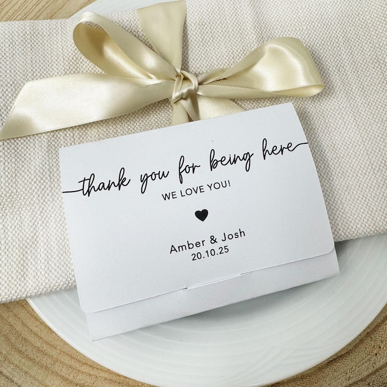 K&ouml;nnte beinhalten: Eine wei&szlig;e Karte mit dem Text "thank you for being here" in schwarzer Schrift, einem schwarzen Herz und den Namen "Amber & Josh" mit dem Datum "20.10.25". Die Karte liegt auf einem wei&szlig;en Teller mit einem beigen Band.