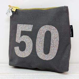 May include: A grey canvas zippered pouch with a sparkly silver number 50 on the front. The pouch has a small crystal charm attached to the zipper.