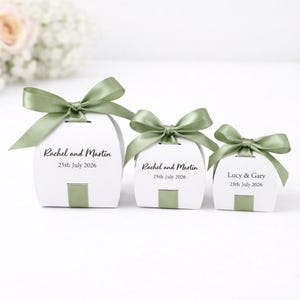 May include: Three white favour boxes of varying sizes, each tied with a sage green satin ribbon bow. The two larger boxes are printed with the text Rachel and Martin 25th July 2026, while the smallest box features the text Lucy and Gary 25th July 2026.