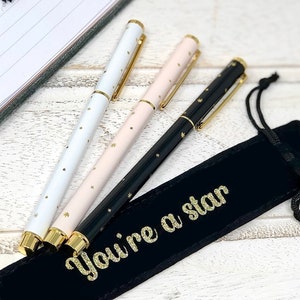 Puede incluir: Tres bolígrafos con punta dorada y detalles de estrellas doradas. Los bolígrafos son blanco, rosa y negro. Los bolígrafos están en una bolsa de terciopelo negro con las palabras "You're a star" escritas en purpurina dorada.