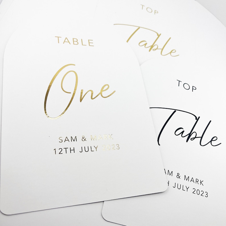 May include: Two white table number cards with gold foil lettering. The top card says "Table One" with the date "12th July 2023" and the names "Sam & Mark". The bottom card says "Table" with the date "1 July 2023" and the names "Im & Mark".