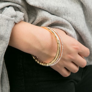 May include: Three gold bangles stacked on a person's wrist. The bangles are hammered and have a textured finish.