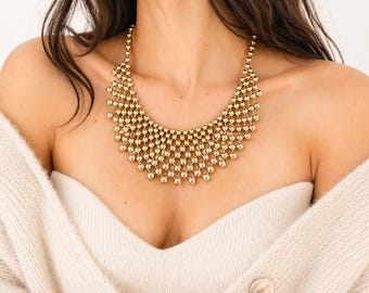 Puede incluir: Un collar dorado llamativo con un diseño de babero. El collar está compuesto por pequeñas cuentas redondas conectadas para formar un patrón similar a una malla. El collar se usa sobre un suéter color crema.