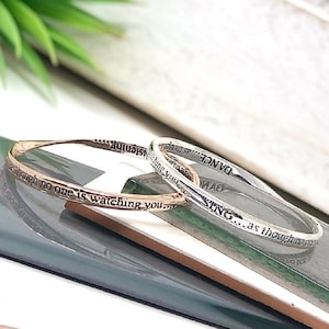 May include: Two silver bangle bracelets with engraved inspirational messages. The top bracelet reads "SING... as though no one is watching you." The bottom bracelet reads "DANCE... as though no one is watching you."