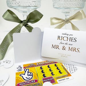 Könnte beinhalten: Eine weiße Karte mit goldenen Lettern, auf der "wishing you RICHES from the new MR. & MRS. STEVEN & AMY 27.09.25" steht. Die Karte ist in der Mitte gefaltet und hat ein Rubbellos daran befestigt.