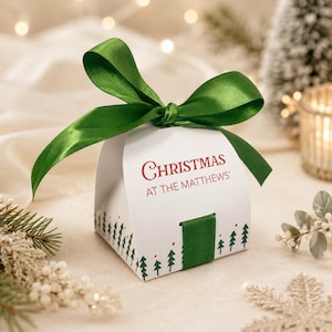 May include: A white, curved gift box featuring a green satin ribbon bow on top. The box is printed with the red text 'CHRISTMAS AT THE MATTHEWS'' and a pattern of small green pine trees along the bottom edge, secured with a green ribbon tab.