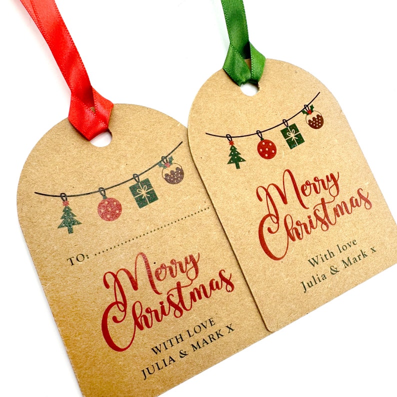 May include: Two brown kraft paper gift tags with red ribbon and green ribbon. The tags feature a string of Christmas decorations, including a Christmas tree, a red bauble, a gift box, and a Christmas pudding. The text on the tags reads "Merry Christmas" with "With love Julia & Mark x" at the bottom.