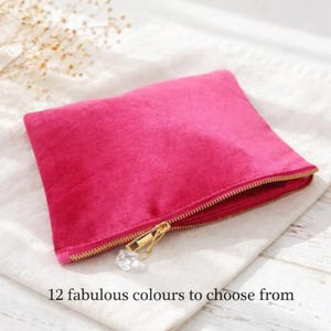 May include: A vibrant fuchsia-colored velvet pouch with a gold zipper and a crystal charm. The pouch is rectangular and rests on a white fabric. Text at the bottom reads "12 fabulous colours to choose from."