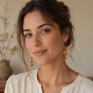 May include: Dangle earrings featuring clusters of small, polished amber and orange gemstone chips suspended from thin gold-toned metal hooks. The earrings hang vertically, showcasing a textured, multi-faceted appearance with warm, earthy tones.