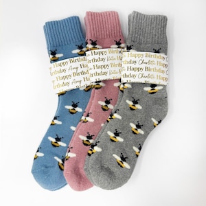 Può includere: Tre paia di calzini blu, rosa e grigi, ciascuno con un motivo a api. I calzini sono avvolti con una fascia che recita "Happy Birthday" e nomi. Un'idea regalo accogliente e allegra.