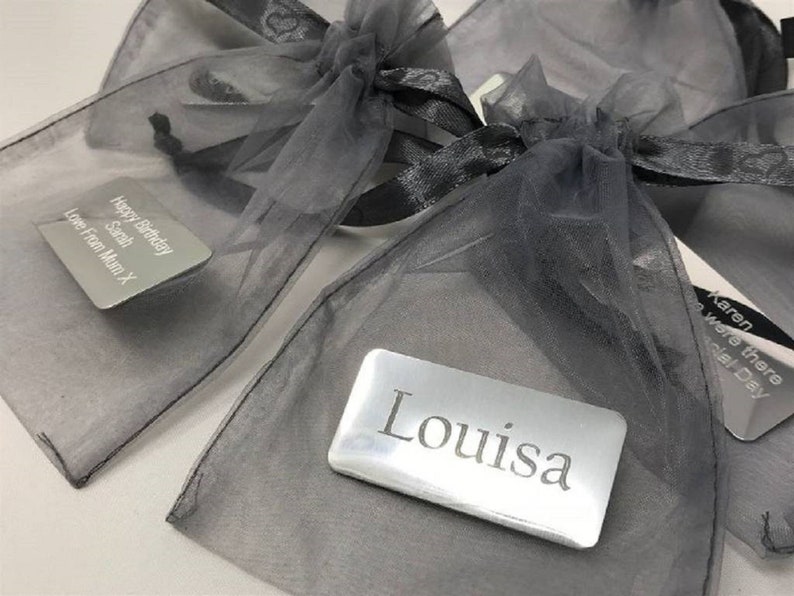 以下が含まれることがあります： 「サラ」と「ルイザ」という名前の入った銀色のギフトタグ2枚が、黒いリボンで結ばれたグレーのオーガンザ製のギフトバッグに入っています。タグには、「ハッピーバースデーサラ」と「ラブフロムマムX」という文字が1枚に、「ルイザ」という文字がもう1枚に書かれています。