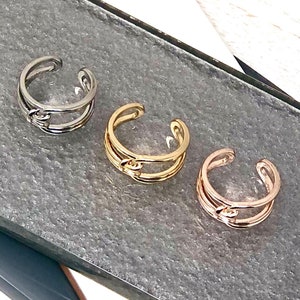 May include: Three gold-toned rings with a knot design. The rings are stacked on a grey surface.