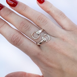 Könnte beinhalten: Ein silberner Ring mit einem Gebiss-Design. Der Ring hat zwei verbundene Schlaufen, die jeweils einem Gebiss ähneln. Der Ring wird an einem Finger getragen und zeigt seinen von Pferden inspirierten Stil. Der Ring besteht aus glänzendem, reflektierendem Metall.