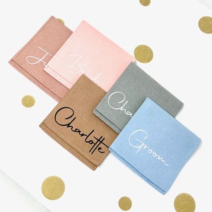 Puede incluir: Cinco pequeñas bolsas cuadradas de fieltro en diferentes colores con nombres escritos en ellas. Las bolsas son rosa claro, marrón, gris, azul claro y rosa oscuro. Los nombres son "Bride", "Charlotte", "Groom", "Cha" y "Ju".