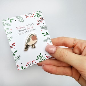 Puede incluir: Un pin de esmalte dorado con un petirrojo con el pecho rojo, posado en una rama. El pin está en una tarjeta blanca con un diseño floral verde y rojo y el texto "Los petirrojos aparecen cuando los seres queridos están cerca".