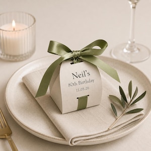 Può includere: Una piccola scatolina portaconfetti curva color bianco sporco, legata con un nastro di raso verde salvia. Sulla scatola è stampato il testo "Neil's 80th Birthday" e la data "11.08.26" con un carattere serif scuro. Il nastro chiude la scatola.