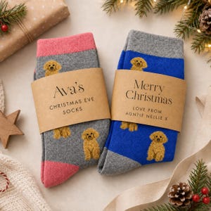 May include: Two pairs of socks with a golden dog pattern. The left pair is grey with pink cuffs and a paper band labeled Ava's Christmas Eve Socks. The right pair is blue with grey cuffs and a paper band labeled Merry Christmas Love From Auntie Nellie X.