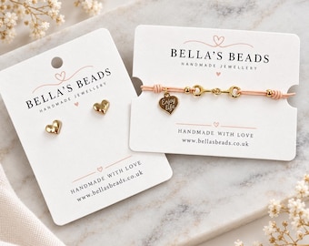 May include: A pair of gold heart-shaped stud earrings and a light pink cord bracelet with a gold-tone bit-style link and a heart charm engraved with the text Enjoy life, both displayed on white branded Bella's Beads jewellery cards.