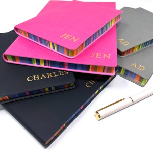 May include: A stack of five personalised notebooks with colourful striped edges. The notebooks are in various colours, including pink, black and grey. The names "Jen", "Charles", and "Ad" are printed in gold on the covers.