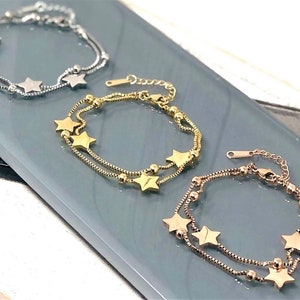 May include: Three delicate layered star charm bracelets in silver, gold, and rose gold. Each bracelet features a chain with small beads and star-shaped charms.
