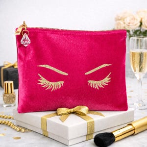 Könnte beinhalten: Eine leuchtend pinke Samttasche mit Reißverschluss und einer goldenen Glitzergrafik von geschlossenen Augen mit langen Wimpern. Der Reißverschlussanhänger ist ein transparenter, diamantförmiger Anhänger. Die Tasche ist rechteckig und weich.