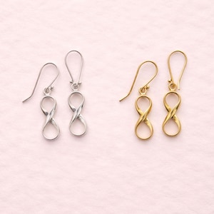 Puede incluir: Dos pares de pendientes, uno plateado y otro dorado, cada uno con un diseño de símbolo de infinito. Los pendientes tienen cierre de gancho y se muestran sobre un fondo rosa suave.