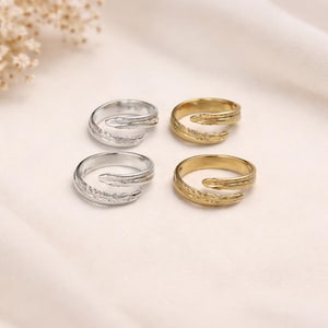 May include: Four rings are shown, two silver and two gold. Each ring features a textured, organic design that wraps around the finger. The rings are set against a soft, off-white background, creating a simple and elegant presentation.