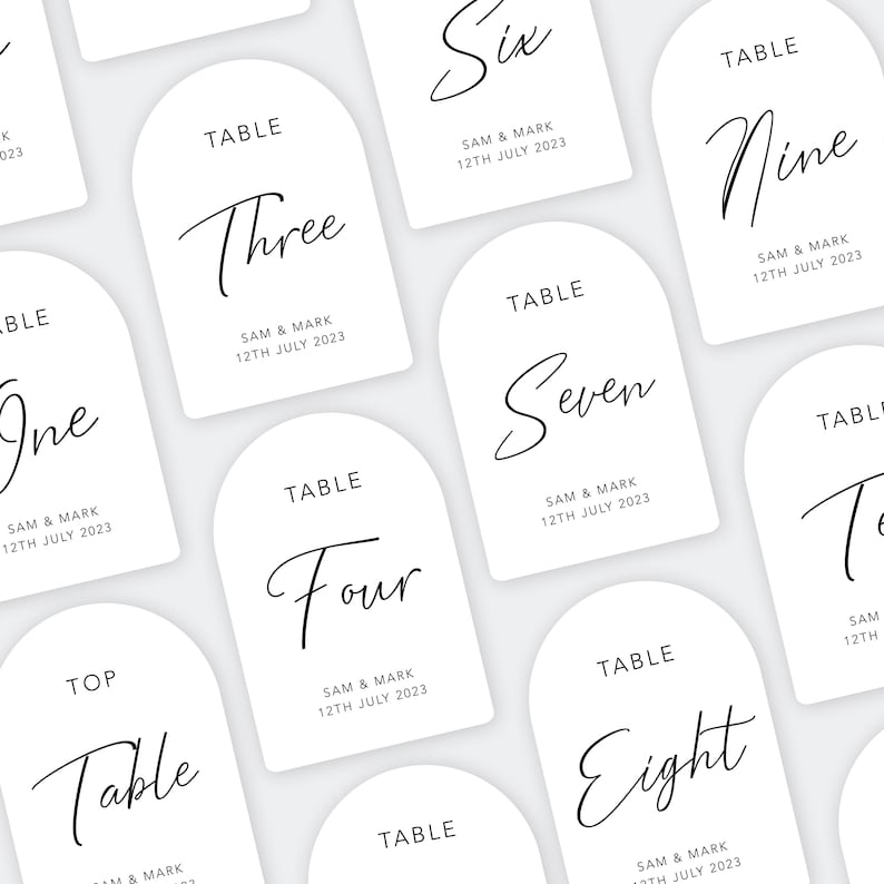 May include: White table number cards with a modern calligraphy font. The cards are arched at the top and have the words "Table" and the table number in black script. The cards also include the names "Sam & Mark" and the date "12th July 2023".